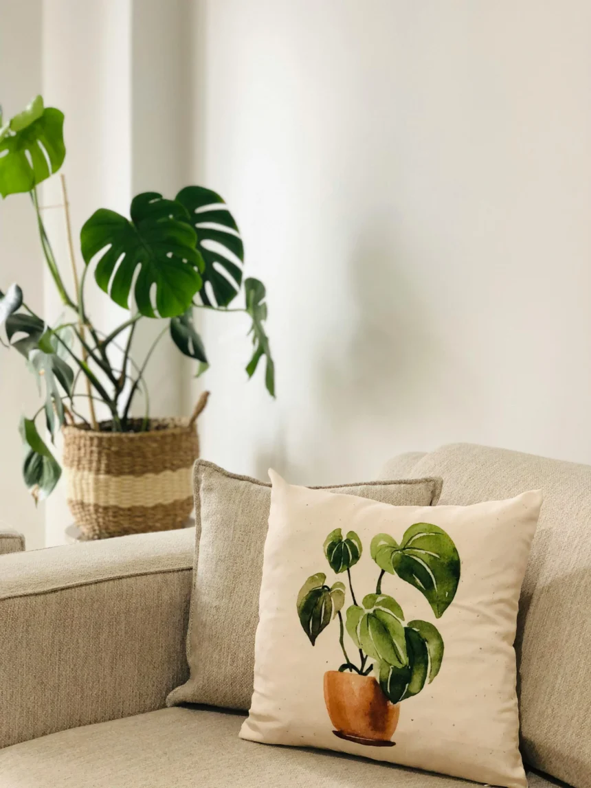 A minimalist living space featuring a beige sofa with a decorative pillow that has a watercolor-style plant illustration, alongside a real monstera plant in a woven basket.