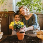 A woman and a young girl planting a flower in a terracotta pot indoors, surrounded by soil and gardening supplies.