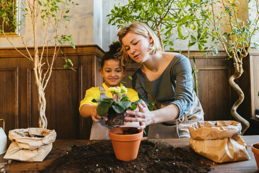 A woman and a young girl planting a flower in a terracotta pot indoors, surrounded by soil and gardening supplies.